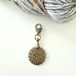 May include: A bronze-colored stitch marker with a floral design and a heart-shaped clasp. The round charm features embossed flowers and leaves. A ball of multicolored yarn is in the background.
