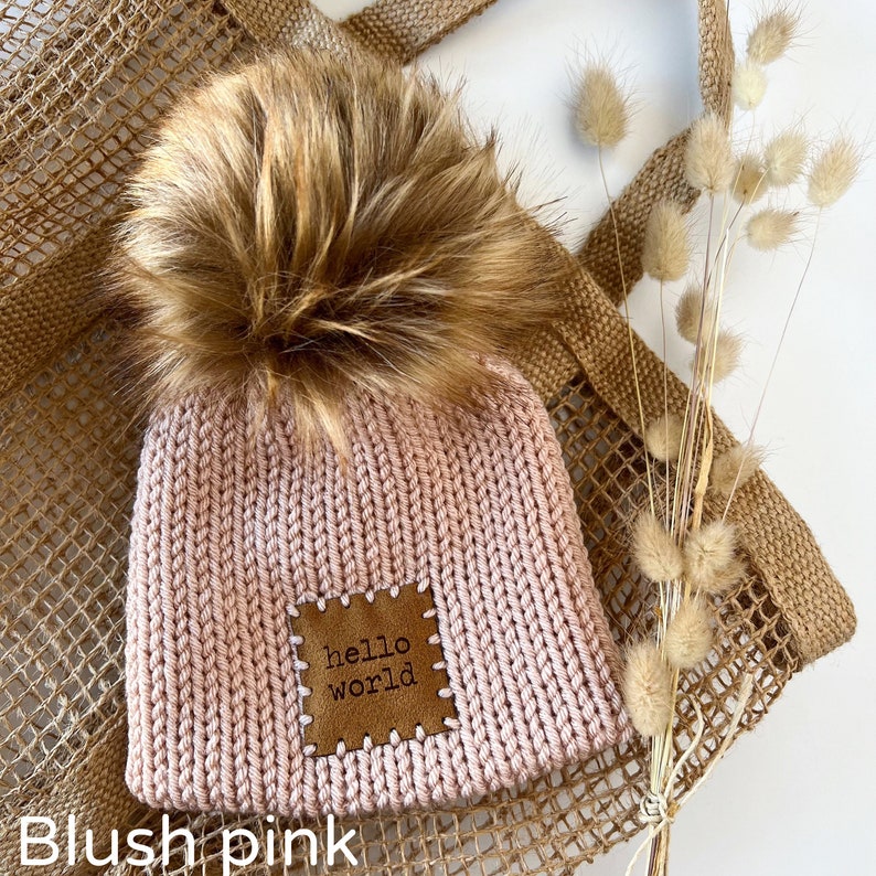Pu&ograve; includere: Berretto lavorato a maglia rosa cipria con un pompon in finta pelliccia. Il cappello presenta una toppa in pelle marrone con le parole "hello world" cucite sopra. Il cappello &egrave; esposto su uno sfondo tessuto.