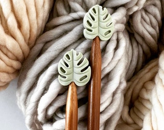 Può includere: Due ferri da maglia in legno sormontati da ciondoli a forma di foglia di monstera verde chiaro. Gli aghi sono posti su uno sfondo di filato spesso e strutturato nei toni del beige e del grigio.
