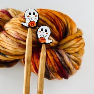 May include: Two wooden knitting needles topped with white ghost charms holding orange jack-o'-lanterns, set against a backdrop of colorful, textured yarn in shades of orange, brown, and red. The needles are made of light-colored wood.