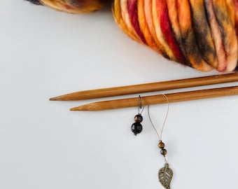Può includere: Primo piano di accessori per maglieria. Un gomitolo di filato colorato nei toni dell'arancione, rosso, giallo e nero è posto sopra due ferri da maglia in legno. Due segnapunti con perline e un ciondolo a forma di foglia sono appesi ai ferri.