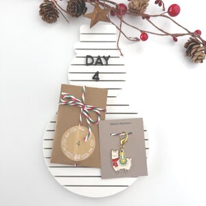 May include: A white snowman-shaped advent calendar with black stripes and the text "DAY 4". It includes a brown paper package tied with red and green twine, and a stitch marker with a llama design. Decorative branches with red berries and pinecones are in the background.