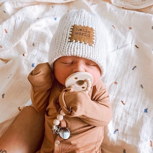 Pu&ograve; includere: Un neonato con un berretto di maglia grigio chiaro con una toppa marrone con la scritta "hello world". Il bambino indossa anche una tutina marrone e ha un ciuccio in bocca. Un fermaciuccio con perline di legno &egrave; attaccato alla tutina.