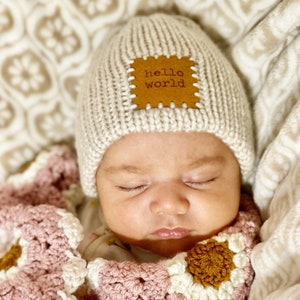 Pu&ograve; includere: Un bambino che dorme con un cappello di maglia beige chiaro con una toppa di cuoio marrone con la scritta "hello world". Il bambino &egrave; avvolto in una coperta all'uncinetto rosa, bianca e marrone.
