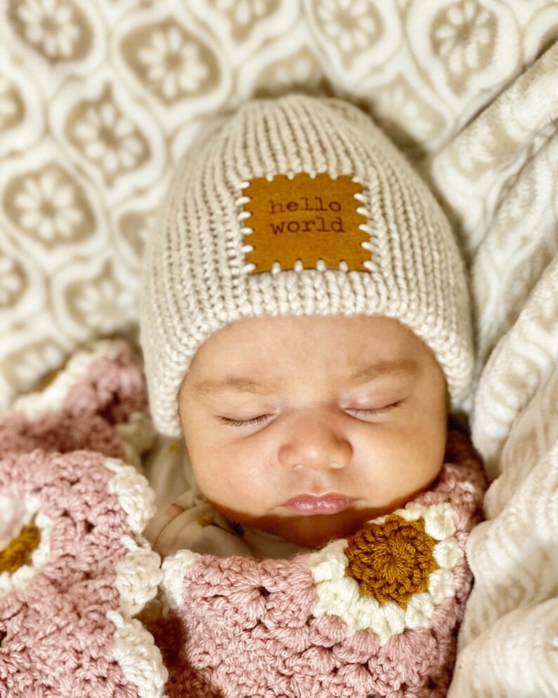 Pu&ograve; includere: Un bambino che dorme con un cappello di maglia beige chiaro con una toppa di cuoio marrone con la scritta "hello world". Il bambino &egrave; avvolto in una coperta all'uncinetto rosa, bianca e marrone.