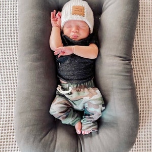 Pu&ograve; includere: Un bambino che dorme con un cappello di lana bianco con una toppa "hello world", una maglietta nera e pantaloni con stampa mimetica. Il bambino &egrave; adagiato in una sdraietta grigia imbottita. Lo sfondo &egrave; una superficie bianca strutturata.