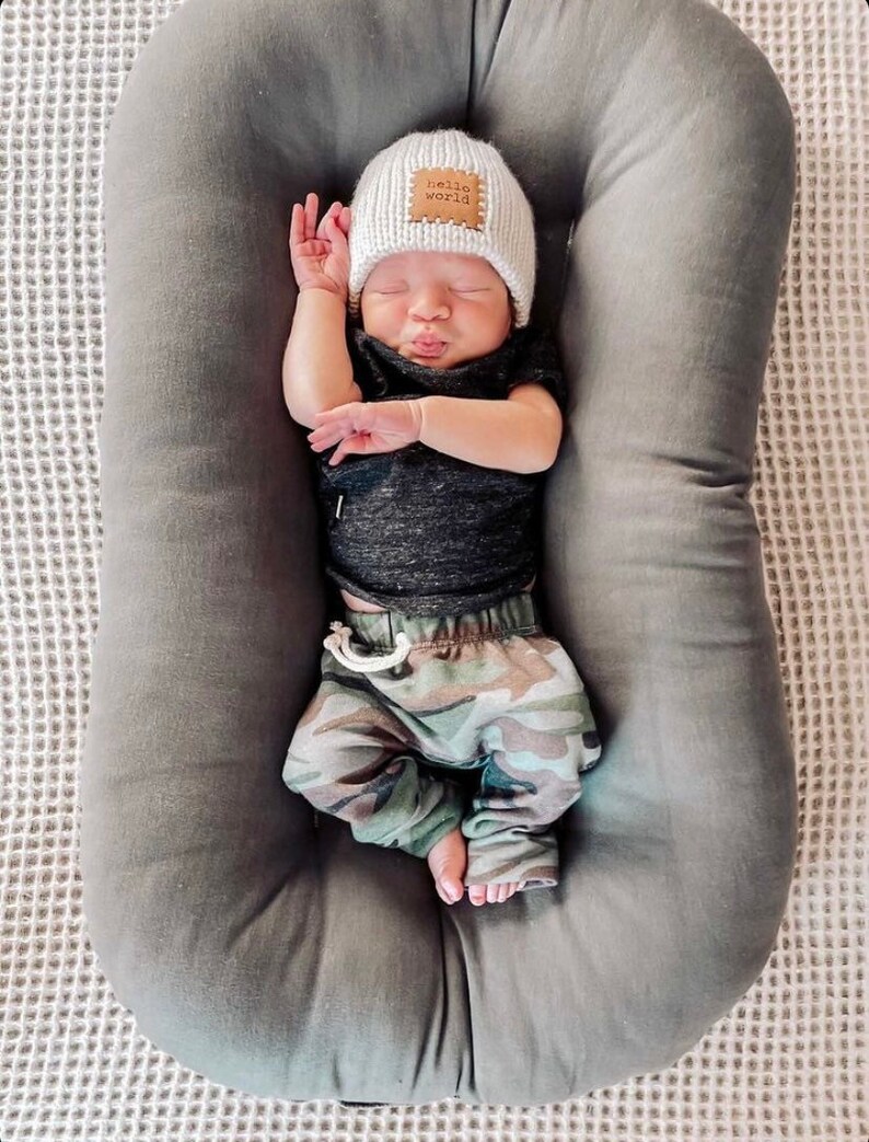 Pu&ograve; includere: Un bambino che dorme con un cappello di lana bianco con una toppa "hello world", una maglietta nera e pantaloni con stampa mimetica. Il bambino &egrave; adagiato in una sdraietta grigia imbottita. Lo sfondo &egrave; una superficie bianca strutturata.
