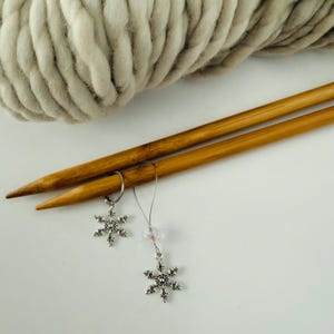 May include: A close-up of knitting supplies. Two wooden knitting needles are positioned diagonally, with two snowflake stitch markers attached. A large ball of light gray yarn is in the background. The needles and markers are on a white surface.