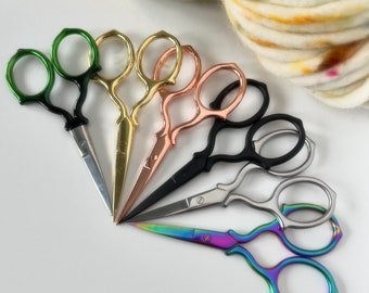 Può includere: Una collezione di piccole forbici da ricamo in vari colori, tra cui verde, oro, oro rosa, nero, argento e arcobaleno iridescente. Le forbici sono disposte a ventaglio, con un gomitolo di lana sullo sfondo.