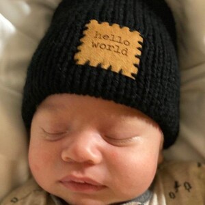 Pu&ograve; includere: Un berretto di maglia nero con una toppa marrone chiaro con la scritta "hello world". Il berretto &egrave; indossato da un bambino. Il bambino indossa una tutina di colore chiaro e sta dormendo.