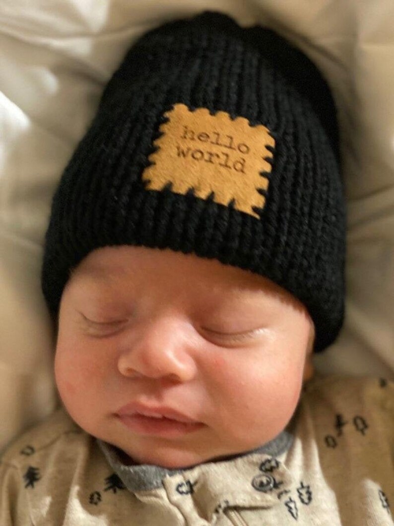 Pu&ograve; includere: Un berretto di maglia nero con una toppa marrone chiaro con la scritta "hello world". Il berretto &egrave; indossato da un bambino. Il bambino indossa una tutina di colore chiaro e sta dormendo.
