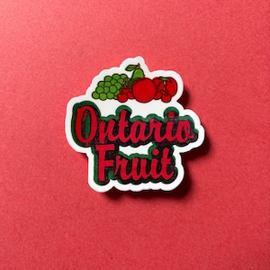 May include: A sticker with the words "Ontario Fruit" in red and green, with a white border. The sticker features illustrations of grapes, apples, and cherries. The background is a solid red color.
