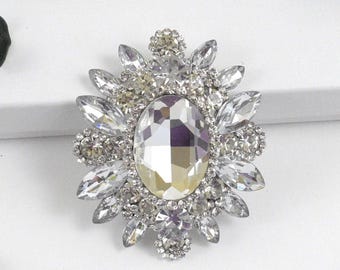 May include: An oval-shaped, silver-toned brooch with a large, faceted, clear central stone. The brooch is surrounded by smaller, clear, sparkling crystals in a floral-like design. The brooch is set against a white background.