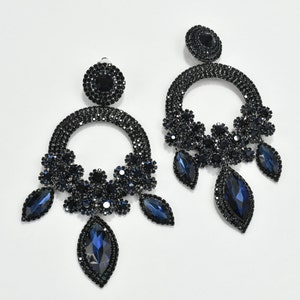 May include: A pair of elegant, dark blue chandelier earrings. Each earring features a circular design encrusted with small, dark blue crystals, with clusters of crystals and teardrop-shaped blue gems hanging below. The earrings are set against a white background.
