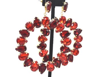 May include: Gold-toned hoop earrings with a double-layered design. Each hoop is adorned with numerous teardrop-shaped, faceted red gemstones. The earrings are displayed on a black stand against a white background.