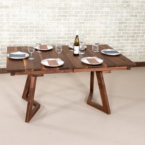 May include: A wooden dining table with a brown finish and a leaf extension. The table has four legs with a unique triangular design. The table is set with four place settings, each with a white plate, a grey napkin, and a wine glass. A bottle of wine is in the centre of the table.