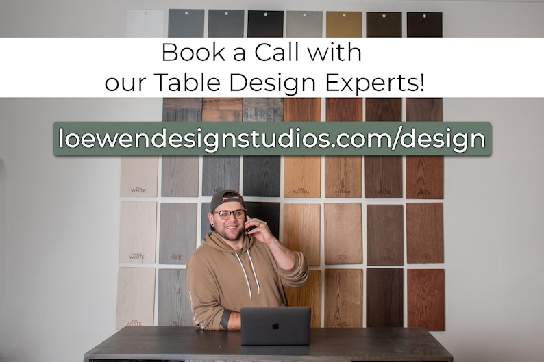 May include: A man in a brown shirt and glasses is on a phone call in front of a wall of wood samples. He is sitting at a table with a laptop in front of him. The text on the wall reads "Book a Call with our Table Design Experts!" and "loewendesignstudios.com/design".