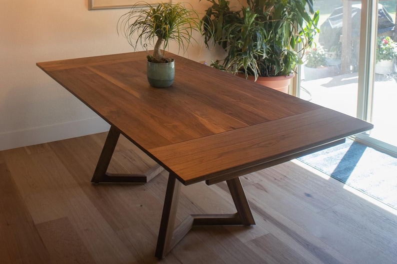 May include: A wooden dining table with a leaf extension, featuring a dark brown wood finish and a simple, modern design. The table has a triangular base and is shown in a room with hardwood floors.