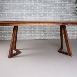 May include: A long, rectangular wooden table with a natural wood finish. The table has a simple, modern design with two triangular legs on each side. The table is made of solid wood and has a smooth, polished surface.