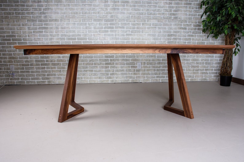 May include: A long, rectangular wooden table with a natural wood finish. The table has a simple, modern design with two triangular legs on each side. The table is made of solid wood and has a smooth, polished surface.