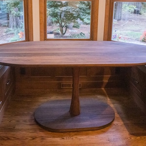 May include: A large oval-shaped wooden table with a pedestal base. The table is made of dark wood and has a natural wood finish. The table is set in a room with a bay window and a built-in bench.