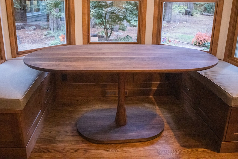 May include: A large oval-shaped wooden table with a pedestal base. The table is made of dark wood and has a natural wood finish. The table is set in a room with a bay window and a built-in bench.
