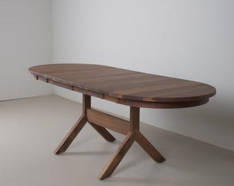 May include: A dark brown wooden oval dining table. The table has a trestle base and is approximately 183 cm long. It is set against a plain white wall and a light grey floor.