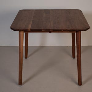 May include: A square wooden table with a dark brown tabletop and four slender, tapered legs. The table has rounded corners and a natural wood grain finish. The legs are a lighter brown color.
