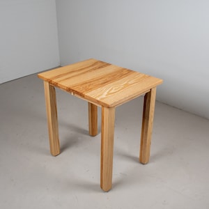 May include: A small wooden table with a light brown finish. The table has a rectangular top and four legs. The table is made of wood with a natural grain pattern.