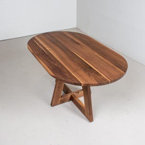 May include: A wooden oval table with a natural wood finish and a simple, modern design. The table has a sturdy base with a cross-shaped support.