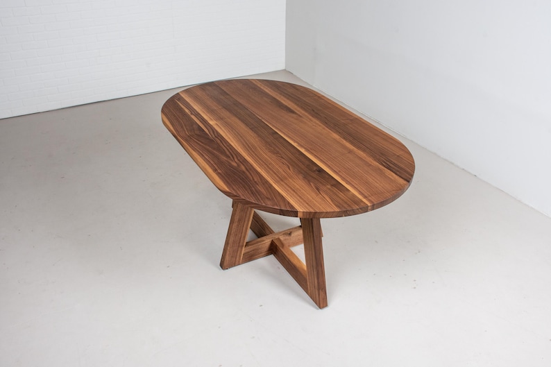 May include: A wooden oval table with a natural wood finish and a simple, modern design. The table has a sturdy base with a cross-shaped support.