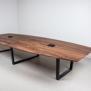 May include: A long, oval-shaped wooden conference table with a dark walnut finish. The table has a black metal frame with four legs. The table is in a white room with a white wall in the background.