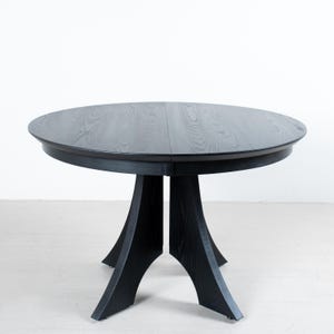 May include: A round, black wooden dining table with a dark finish. The table has a circular top and a pedestal base with four angled legs. The table is approximately 48 inches in diameter.