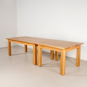 May include: A long rectangular wooden table with a natural light brown finish, featuring a smooth flat top surface and four sturdy square legs at each corner, constructed from solid wood with visible grain patterns throughout the entire structure.