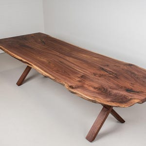 May include: A large, rustic wooden dining table with a live edge top and a natural wood finish. The table has a unique X-shaped base made of the same wood.
