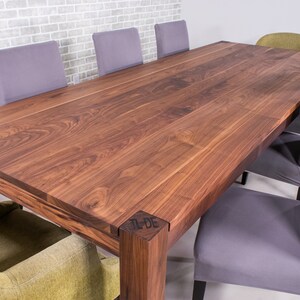 Peut inclure: Une grande table à manger rectangulaire en bois avec une finition brun foncé. La table a un design simple avec des pieds droits et une surface lisse. La table est entourée de chaises avec un revêtement gris.