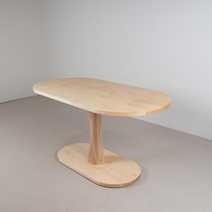 May include: A light-coloured wooden table with an oval top and base, connected by a central, curved support. The table is made of unfinished wood, showcasing its natural grain. The table's dimensions are approximately 122 cm long and 61 cm wide.