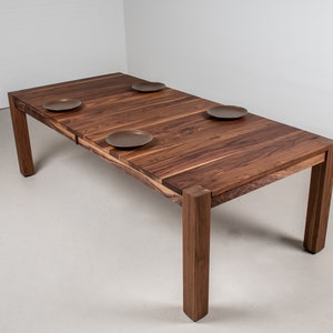 May include: A dark brown wooden dining table with a rectangular top and four legs. The table has a leaf extension that can be added to increase the length. There are four brown ceramic plates on the table.
