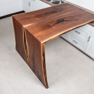 May include: A large, live edge walnut wood countertop with a unique split in the center. The countertop is mounted on a white kitchen cabinet.