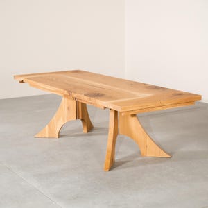 May include: A light-colored wooden dining table with a rectangular top and unique, curved legs. The table appears to be made of solid wood, showcasing natural grain patterns. The design is modern and minimalist, suitable for a dining room or kitchen.