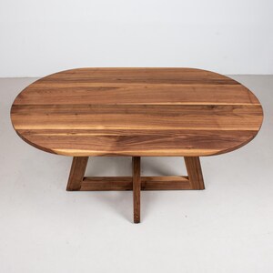 May include: A wooden oval table with a natural wood finish and a simple, modern design. The table has a cross-shaped base.