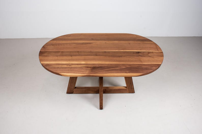 May include: A wooden oval table with a natural wood finish and a simple, modern design. The table has a cross-shaped base.