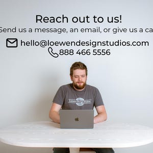 May include: A person sitting at a white table, using a laptop computer. The text on the wall reads "Reach out to us! Send us a message, an email, or give us a call! hello@loewendesignstudios.com 888 466 5556"