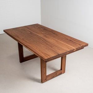 May include: A rectangular wooden dining table with a dark brown finish. The table has a simple, modern design with a wide, flat top and a square base.