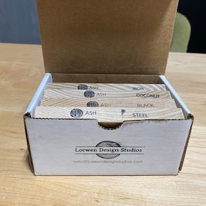 May include: A white box with the words "Loewen Design Studios" and "hello@loewendesignstudios.com" printed on the front. The box is open and contains several wooden blocks with different wood types and finishes labeled on each block. The wood types include Ash, Iron, Coconut, Black, and Steel.