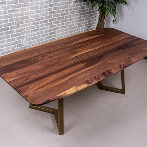 May include: A wooden dining table with a natural walnut finish and a curved edge. The table has a gold metal base with a geometric design.