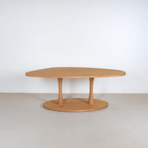 May include: A light brown wooden table with an oval top and two cylindrical legs. The legs connect to a rounded base. The table is set against a white wall and a light grey floor.