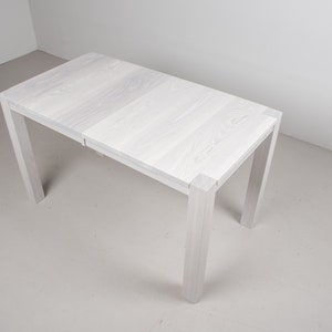 May include: White wooden table with a simple design. The table has a rectangular top and four legs. The table is made of solid wood and has a natural finish.