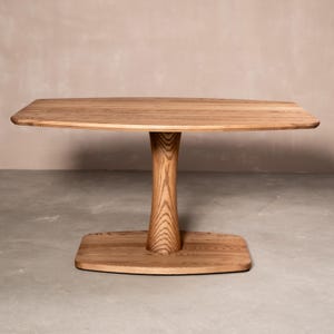 May include: A solid wood table with a rectangular top and a unique pedestal base. The table is made of light-colored wood, showcasing the natural grain. The base is a sculpted, hourglass shape, and the table is suitable for dining or as a decorative piece.