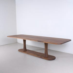 May include: A long, rectangular wooden table with a natural finish. The table is supported by two curved, pedestal-style legs connected by a lower stretcher. The table's dimensions are approximately 96 inches long, 36 inches wide, and 30 inches high.